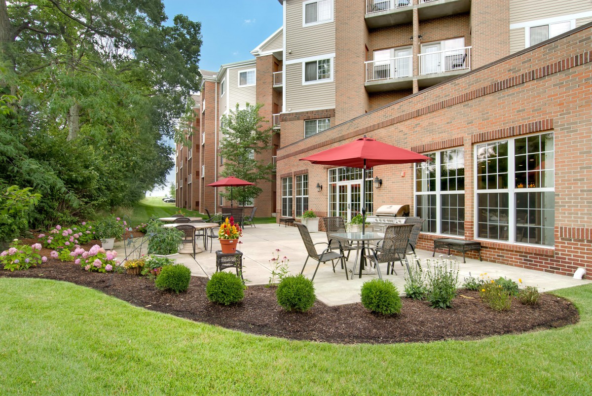A well-maintained outdoor patio area with tables, chairs, and red umbrellas in front of an apartment building.