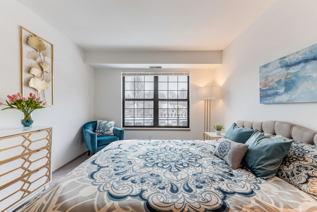 A cozy bedroom with a large window, featuring a stylish bedspread and a comfortable chair.