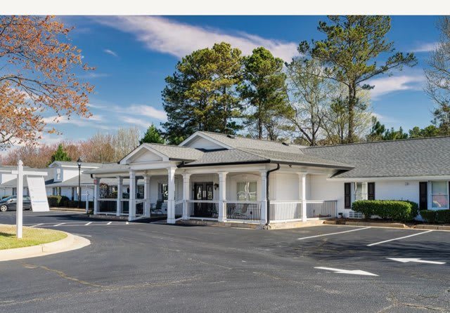 A white building with columns and a parking lot surrounded by trees.