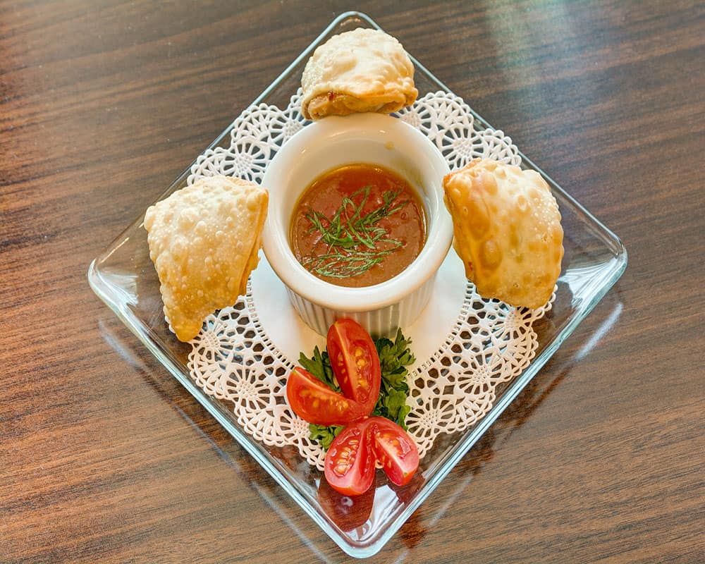 A beautifully presented appetizer featuring samosas served with a dipping sauce, garnished with tomato slices and parsley.