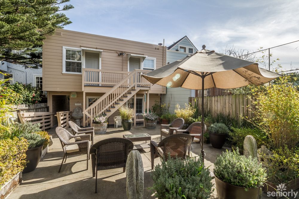 A cozy backyard patio with ample seating, surrounded by lush plants and shaded by an umbrella.