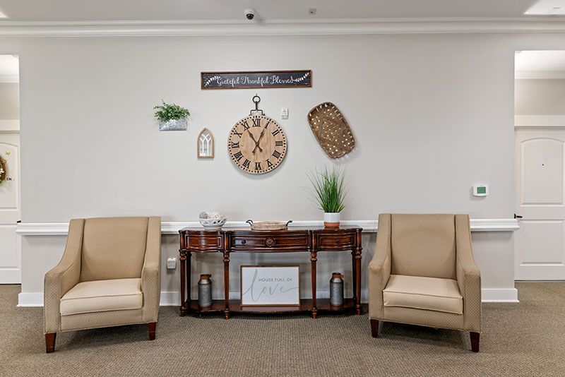 A cozy living room seating area with two beige armchairs, a console table, and decorative wall elements including a clock and plants.