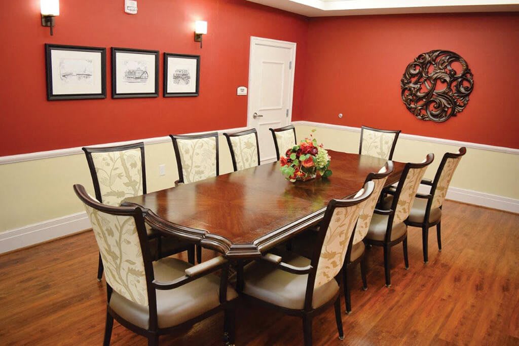 A formal dining room with a long wooden table and elegant chairs, surrounded by artwork and warm red walls.