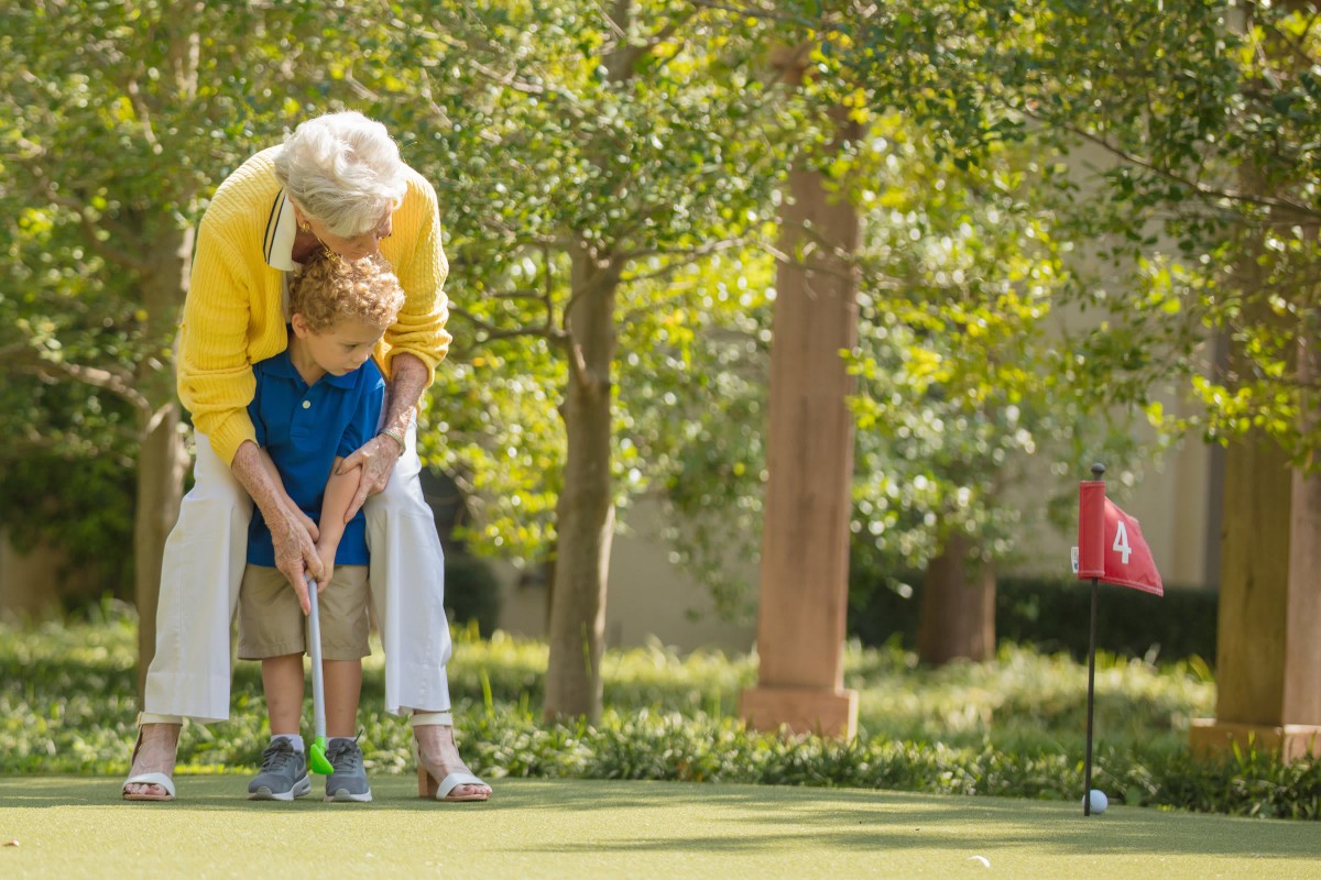 A senior person helps a child play golf outdoors on a sunny day.