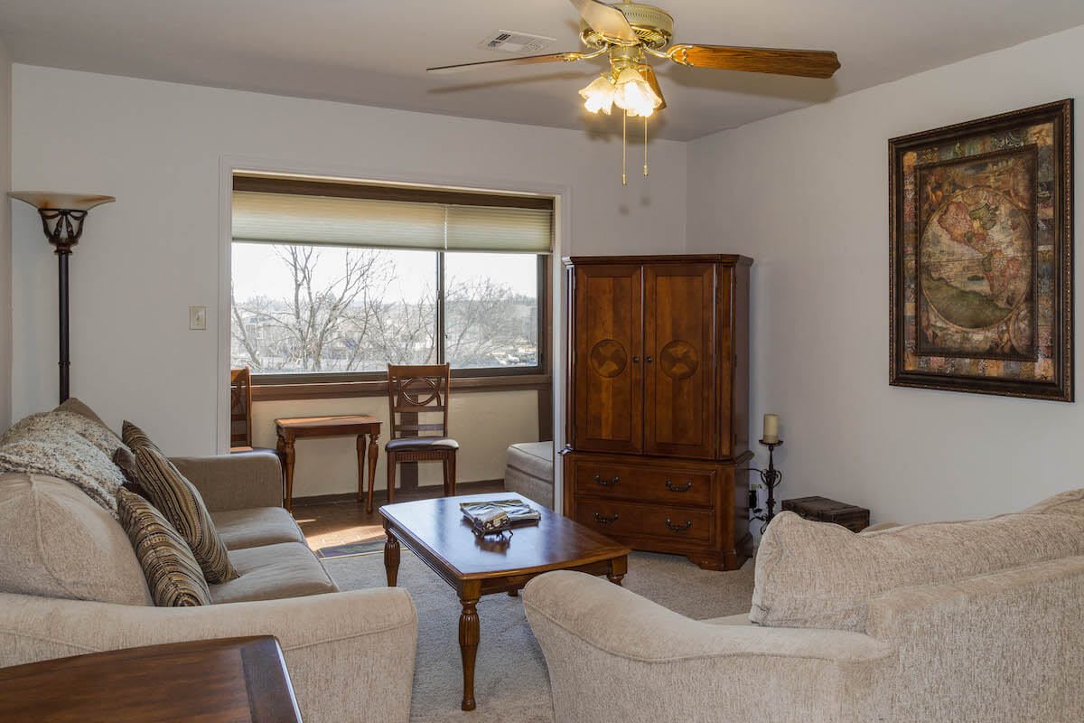 A cozy living room featuring a sofa, armchair, coffee table, and wooden cabinet. Natural light streams in through the large window.