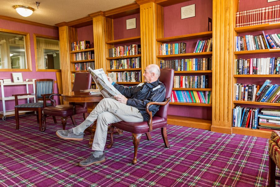 A person sits comfortably in a library, reading a newspaper amidst rows of bookshelves.
