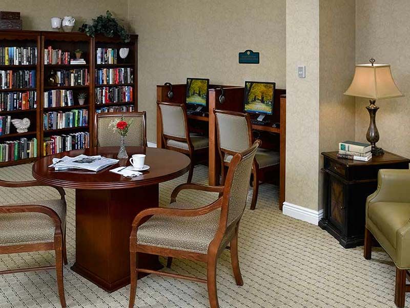 A cozy library room with a bookshelf, round table, chairs, and computer stations.