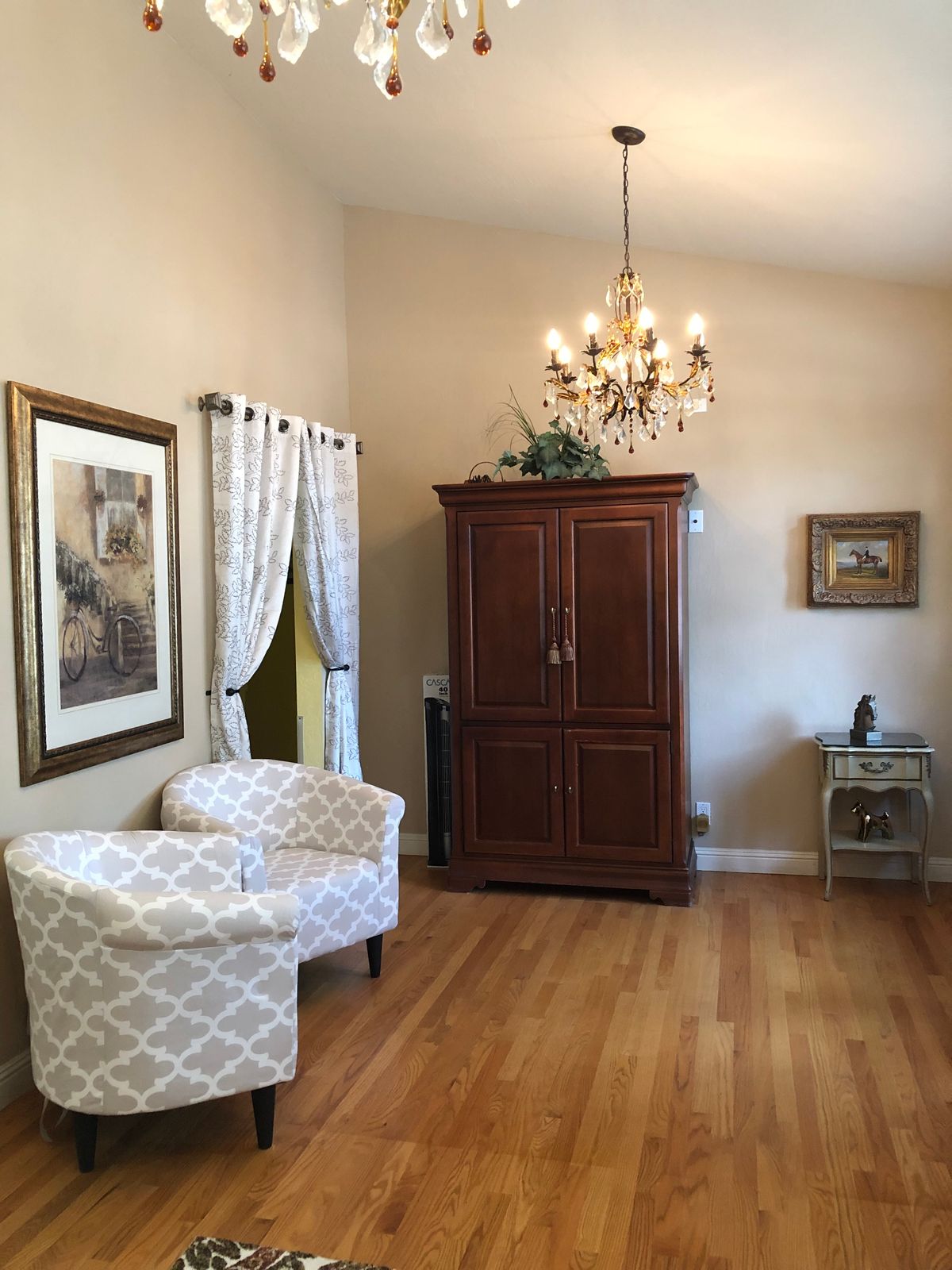 A cozy living room with two patterned armchairs, a wooden cabinet, a chandelier, and hardwood flooring.