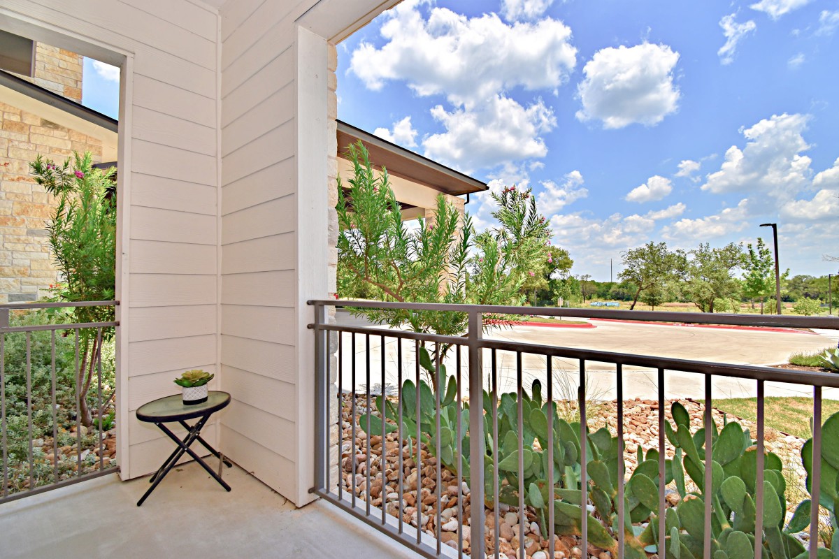 A small outdoor balcony with a cactus and a table under a bright blue sky with fluffy clouds.