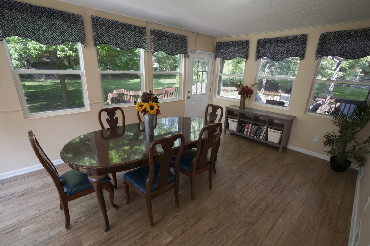 A bright dining room with a wooden table and chairs, adorned with a vase of sunflowers and surrounded by large windows.