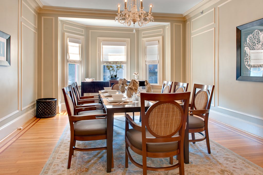 An elegant dining room with a long table and chandelier, surrounded by wooden chairs and large windows.
