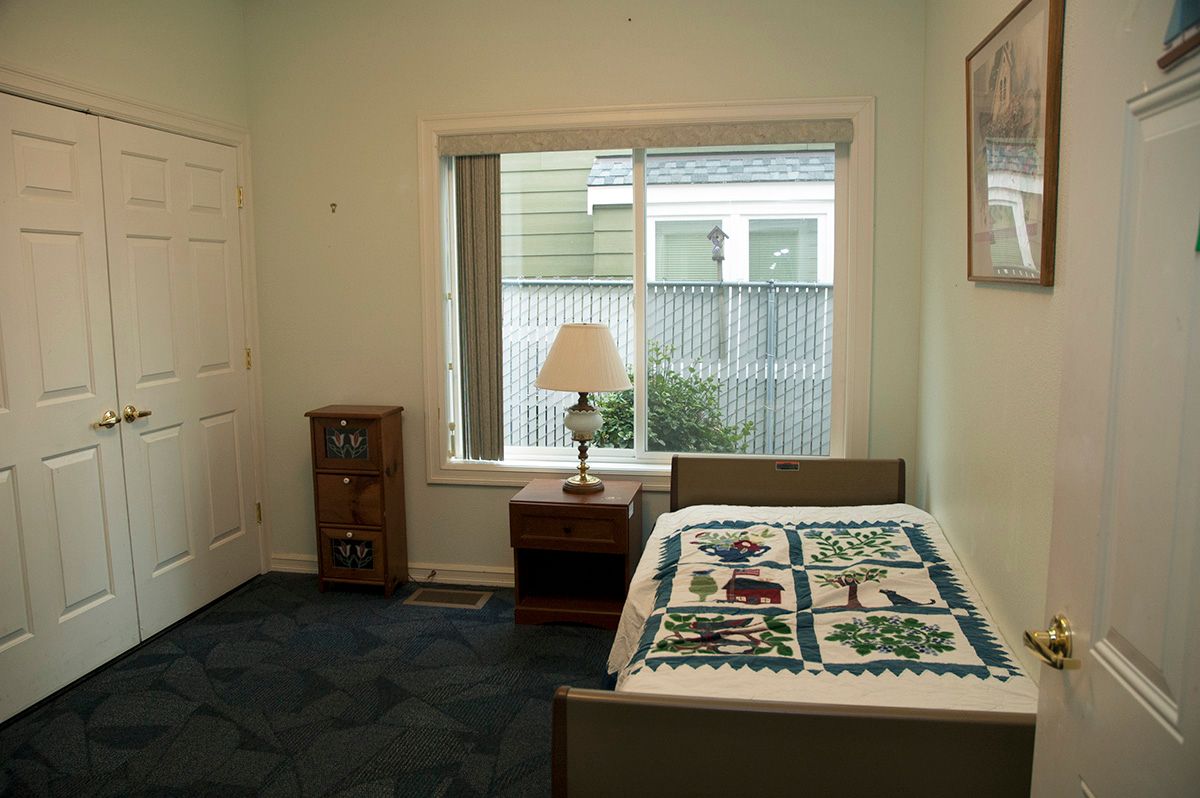 A simple bedroom with a single bed, quilted blanket, nightstand, and a lamp by the window.