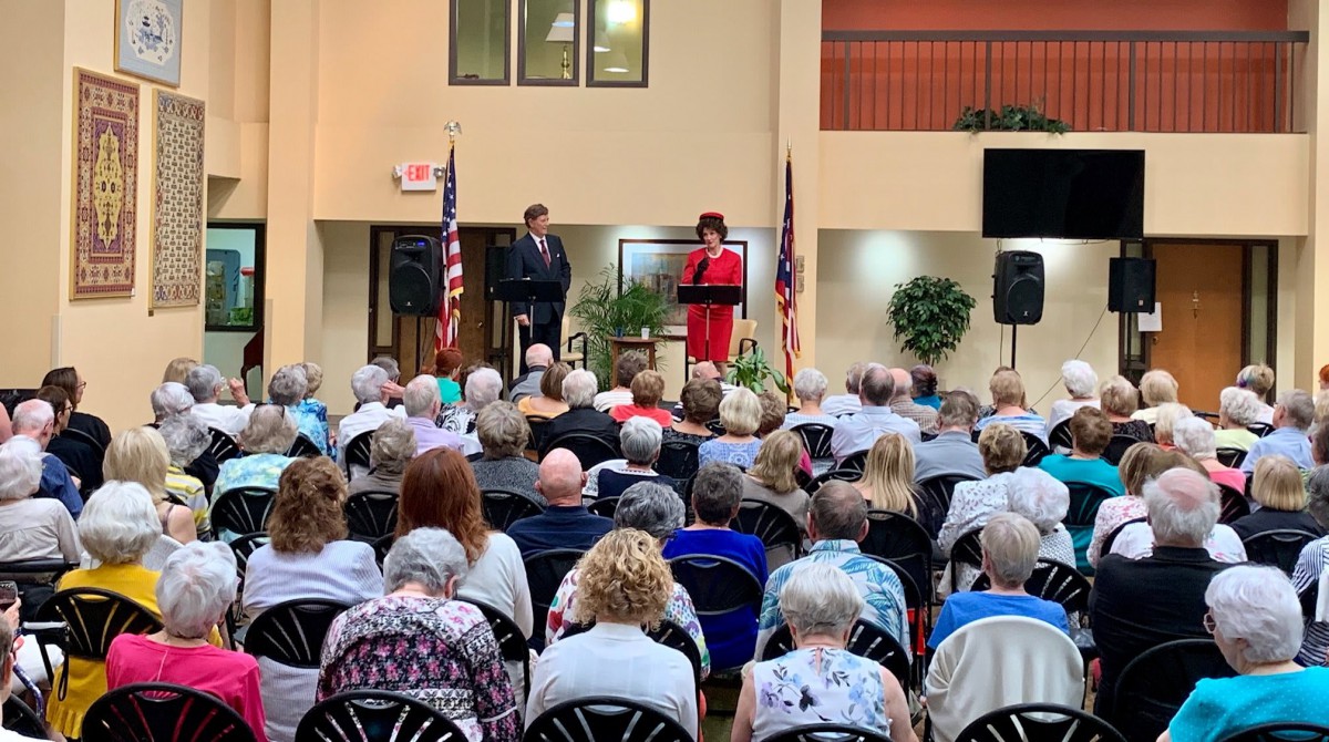 Two speakers addressing an audience in an indoor setting with flags displayed.
