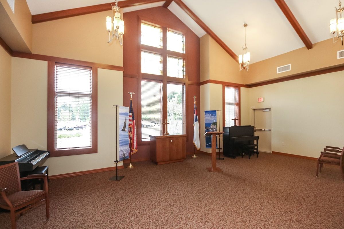 A well-lit room with large windows, carpeted flooring, and various pieces of furniture.
