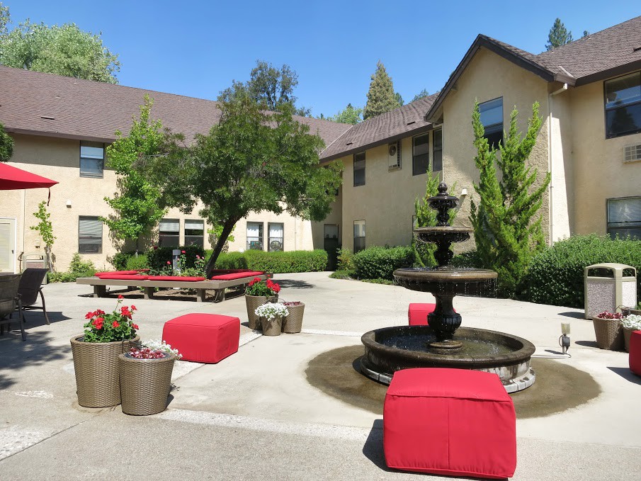 A sunny courtyard with red outdoor furniture and a central fountain, surrounded by buildings and greenery.