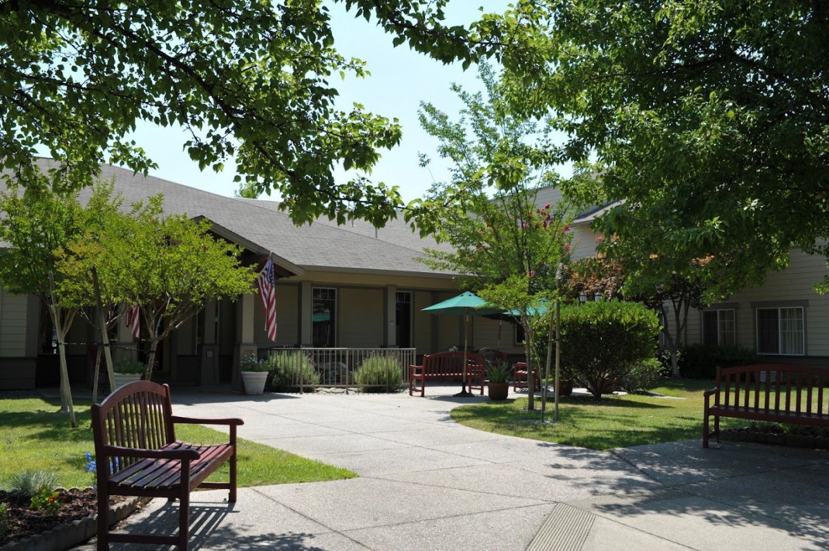 A cozy house surrounded by trees and a garden, featuring benches and a patio.