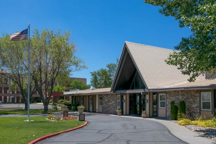 A modern building with a triangular roof, surrounded by trees and a paved driveway under a clear blue sky.