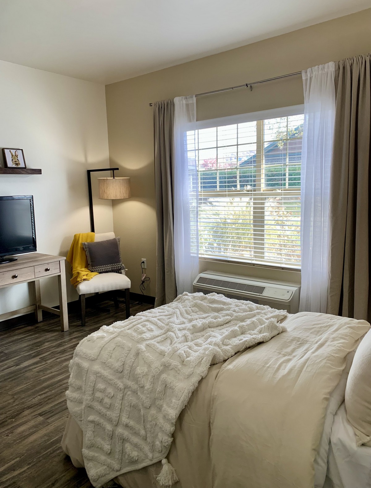A cozy bedroom interior featuring a neatly made bed, a lamp, and a desk with a TV near a large window.