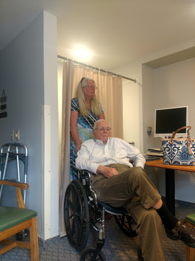 A woman stands behind an elderly man in a wheelchair, who is seated in a well-lit room with a computer nearby.