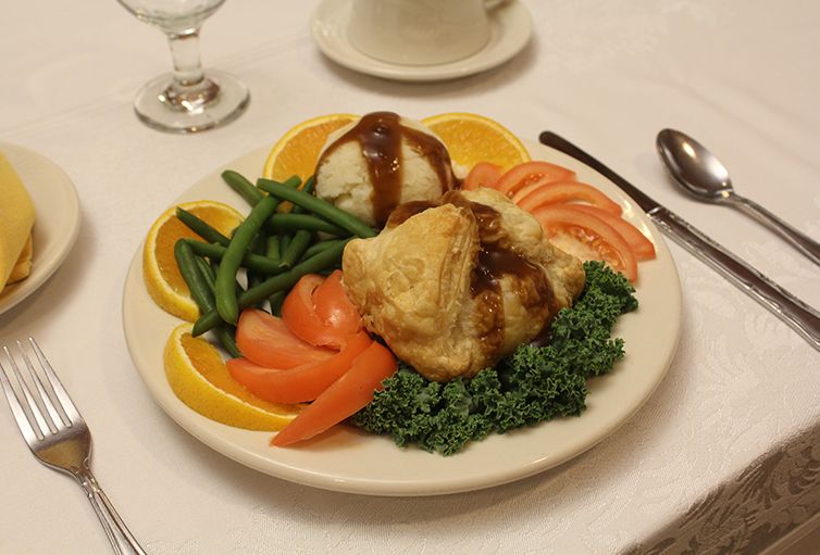 A plated dinner featuring roasted chicken, mashed potatoes topped with gravy, green beans, sliced tomatoes, and orange slices garnished with parsley.