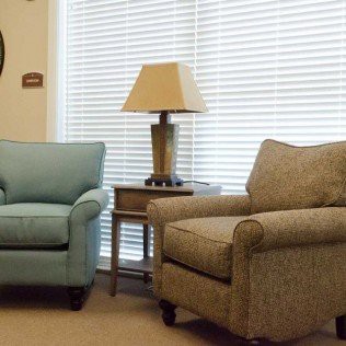 A cozy living room setting with two armchairs and a lamp on a side table, complemented by window blinds in the background.