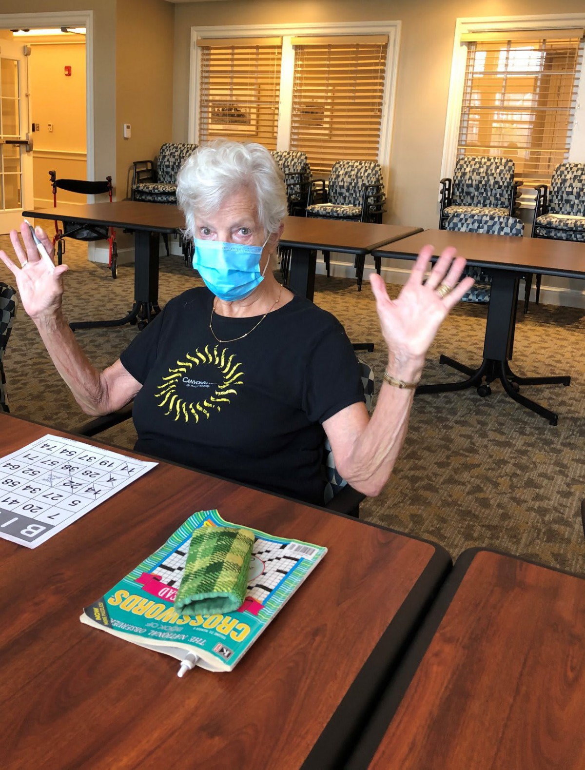 A senior woman wearing a mask is playing bingo indoors with hands raised.
