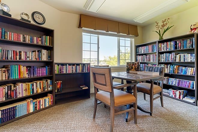 A cozy library reading room with bookshelves, a table, and chairs by a large window.