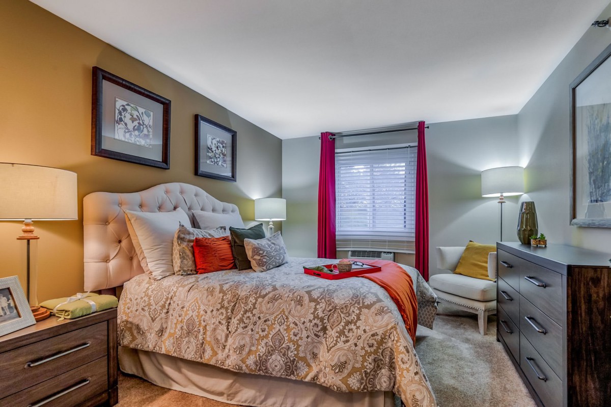A cozy and stylishly decorated bedroom featuring a plush bed with patterned bedding, framed artwork on the walls, and vibrant red curtains framing a window.