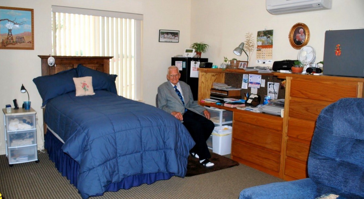 An elderly man sitting at a desk in a bedroom with a blue bedspread.