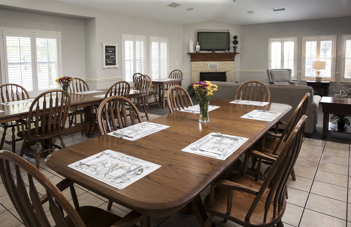 A clean and well-organized dining room with wooden tables and chairs, set with placemats and decorated with a vase of flowers.