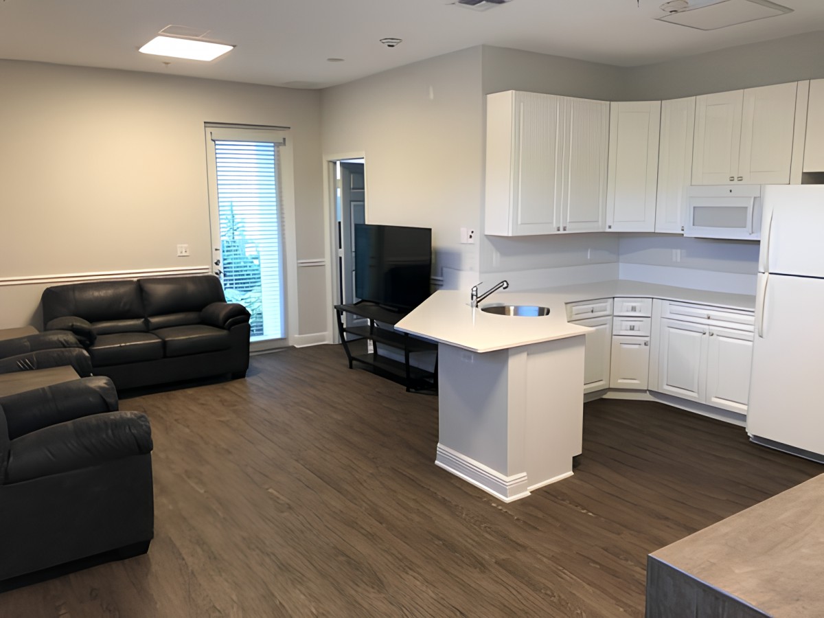 A modern open-concept living room and kitchen with dark couches, a TV, and hardwood floors.