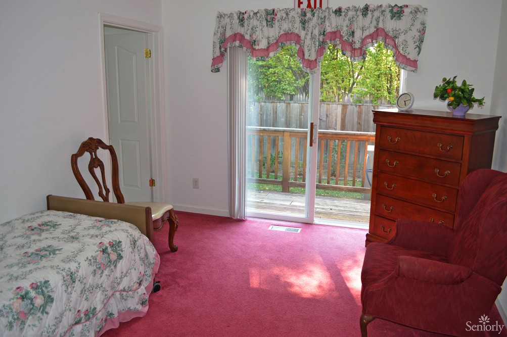 A cozy bedroom with pink carpet and classic furniture, featuring a large window with floral curtains that opens to a view of the garden.