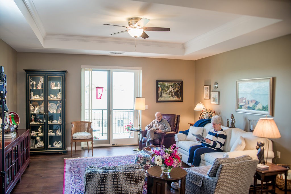 A cozy living room with two people relaxing on sofas, surrounded by home decor and illuminated by natural light from a large window.