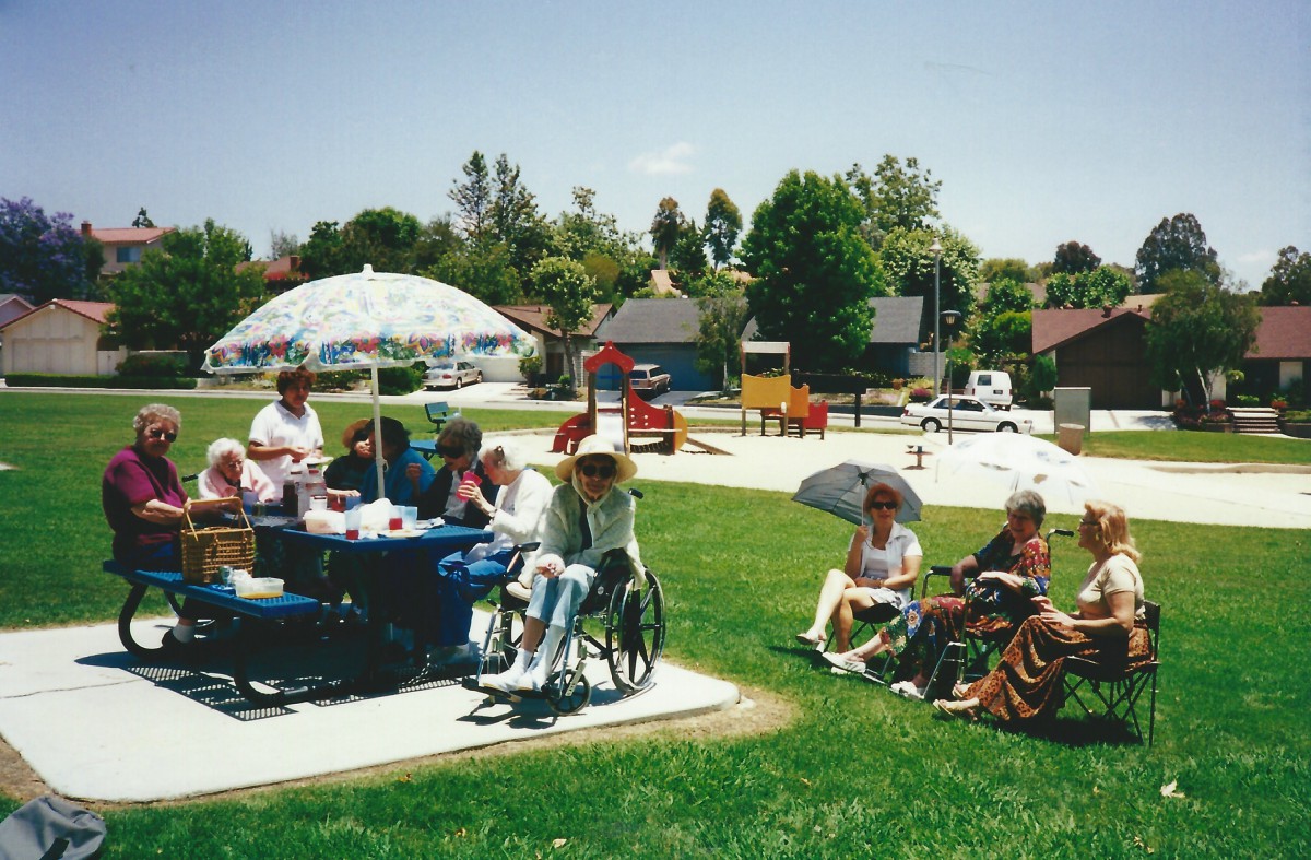 A group of people enjoying a sunny day at a park, sitting together under a large umbrella with some in wheelchairs.