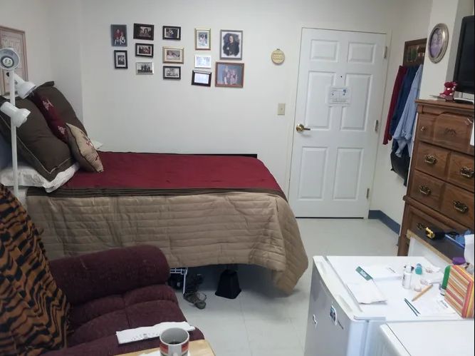 A small bedroom with a bed, chair, and fridge, featuring pictures on the wall.