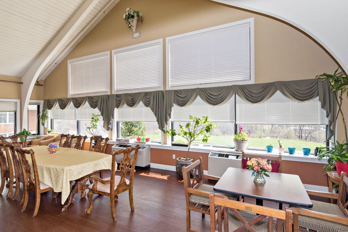 A well-lit dining room featuring wooden tables and chairs, with large windows adorned with blinds and curtains.