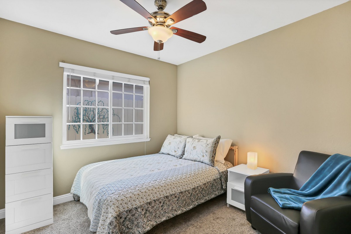 A cozy bedroom with a bed, nightstand, and armchair, featuring a ceiling fan and large window.