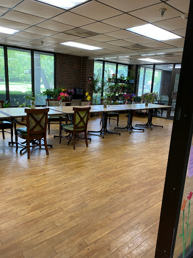 A spacious and well-lit office workspace featuring multiple tables and chairs, surrounded by large windows and greenery.