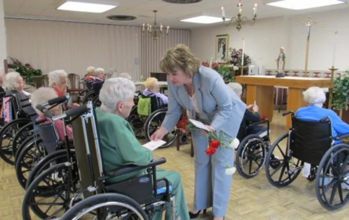 A woman interacts with seniors in wheelchairs at a nursing home, offering them flowers.