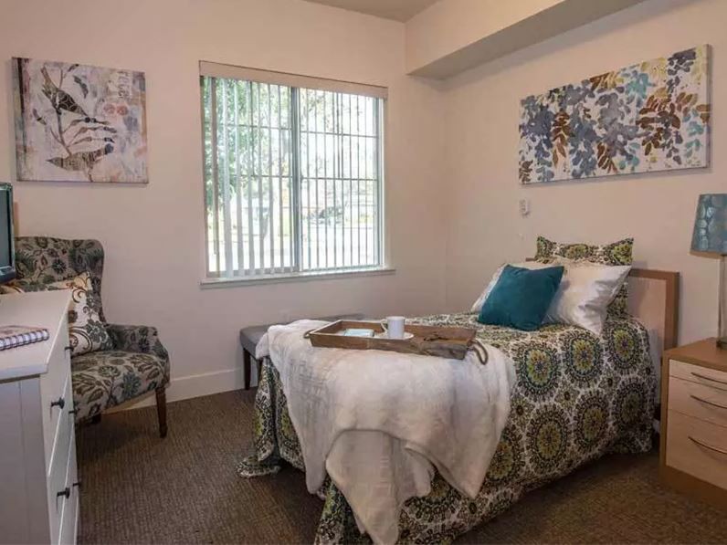 A cozy bedroom with a single bed, decorative pillows, a patterned chair, and artwork on the walls.