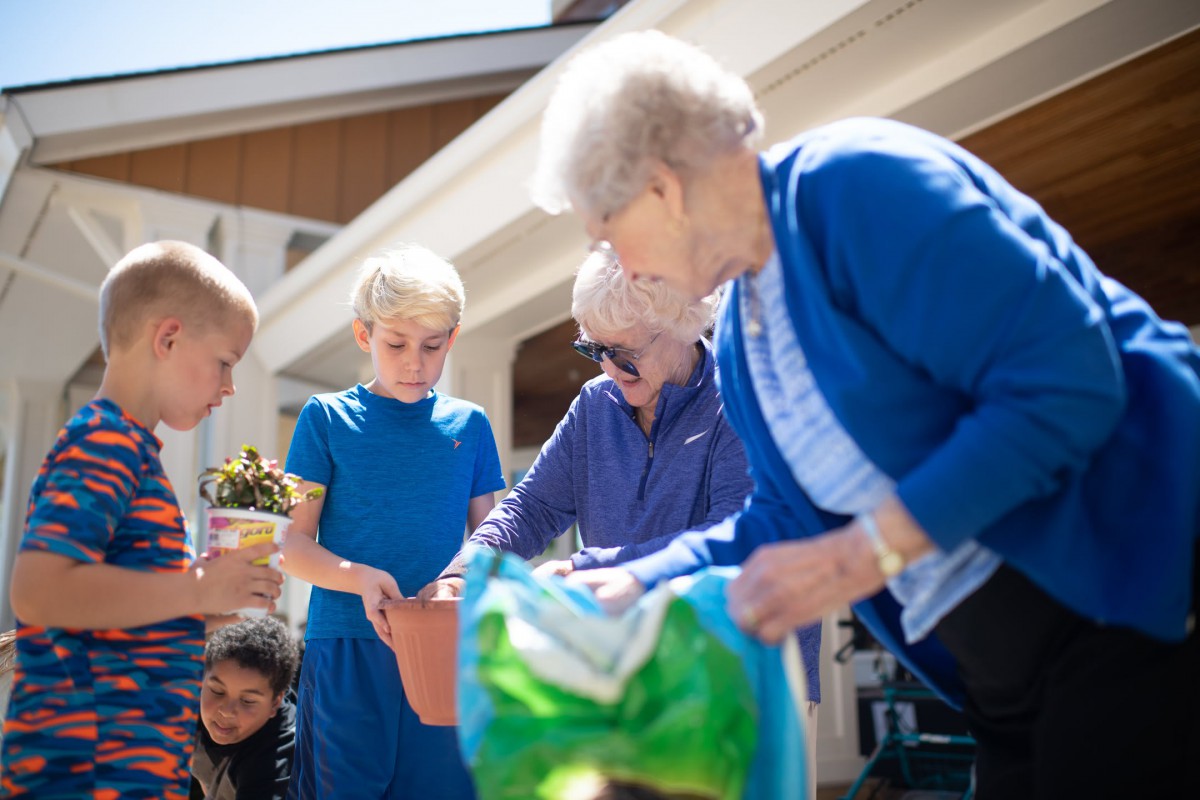 A group of people, including an elderly woman and children, engage in gardening activity outdoors.