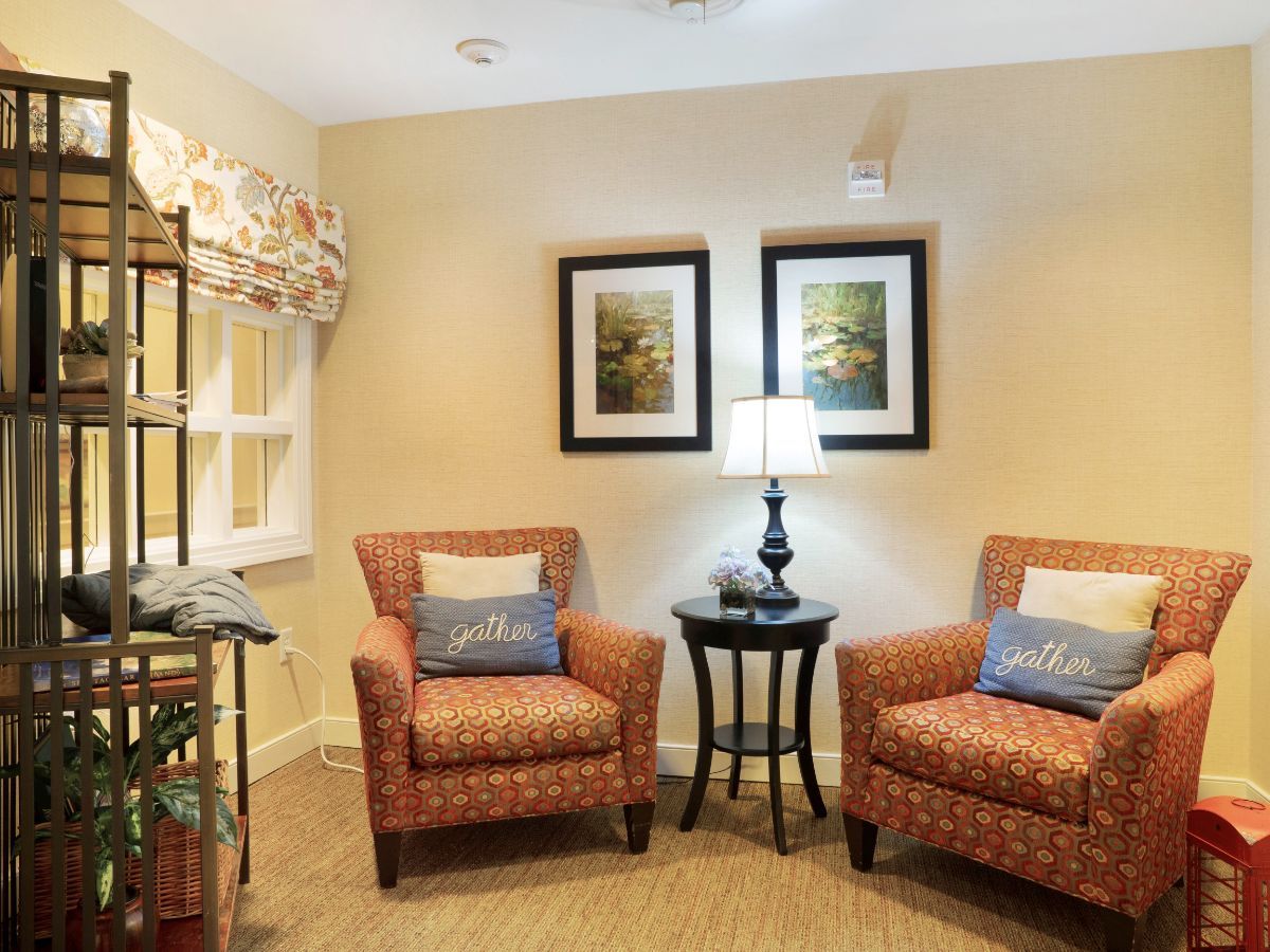 A cozy living room corner with two patterned armchairs, a lamp on a small table, and framed artworks adorning the wall.