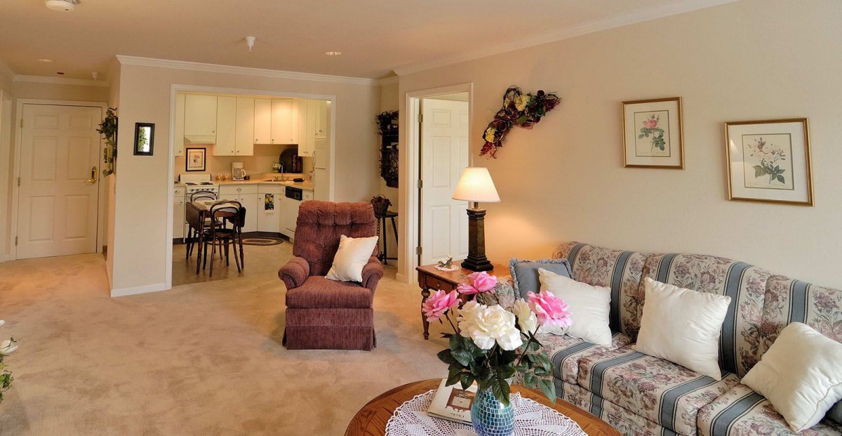 A cozy living room with a brown armchair, patterned sofa, and a flower arrangement on the table. The space opens into a kitchen area.