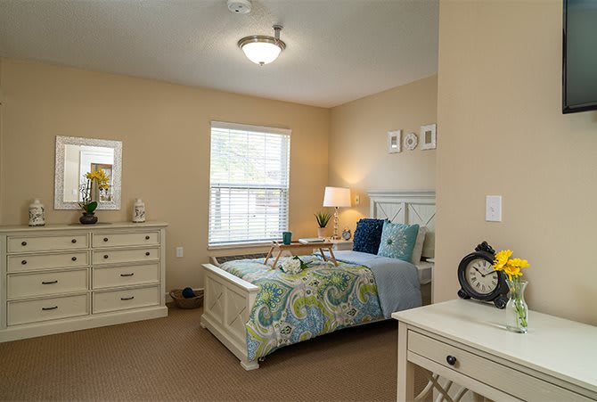 A cozy bedroom featuring a bed with colorful bedding, a dresser, and a lamp by the window.