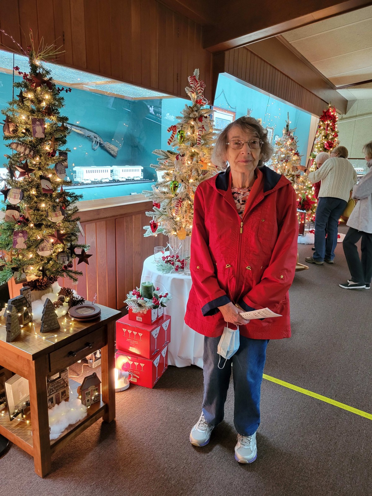 A woman in a red coat stands among festive Christmas decorations and trees indoors.
