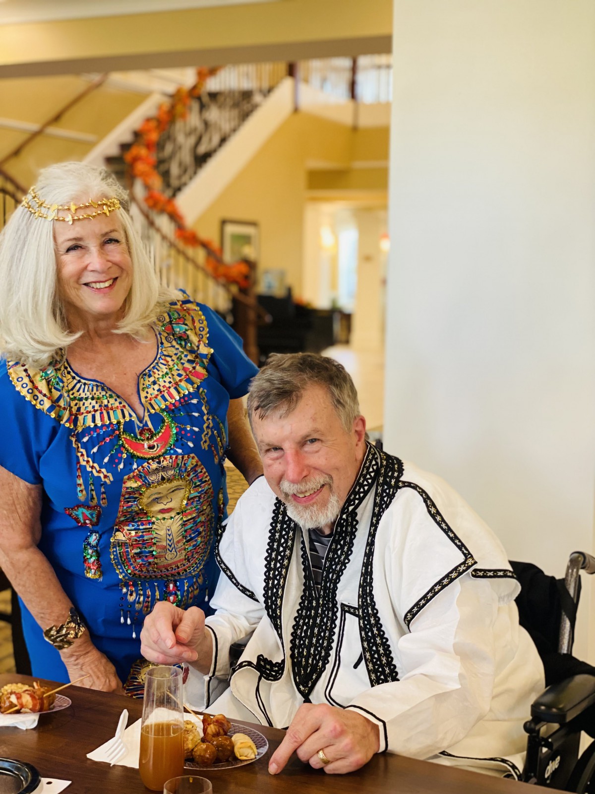 An elderly couple in festive, cultural costumes indoors, smiling for the camera.