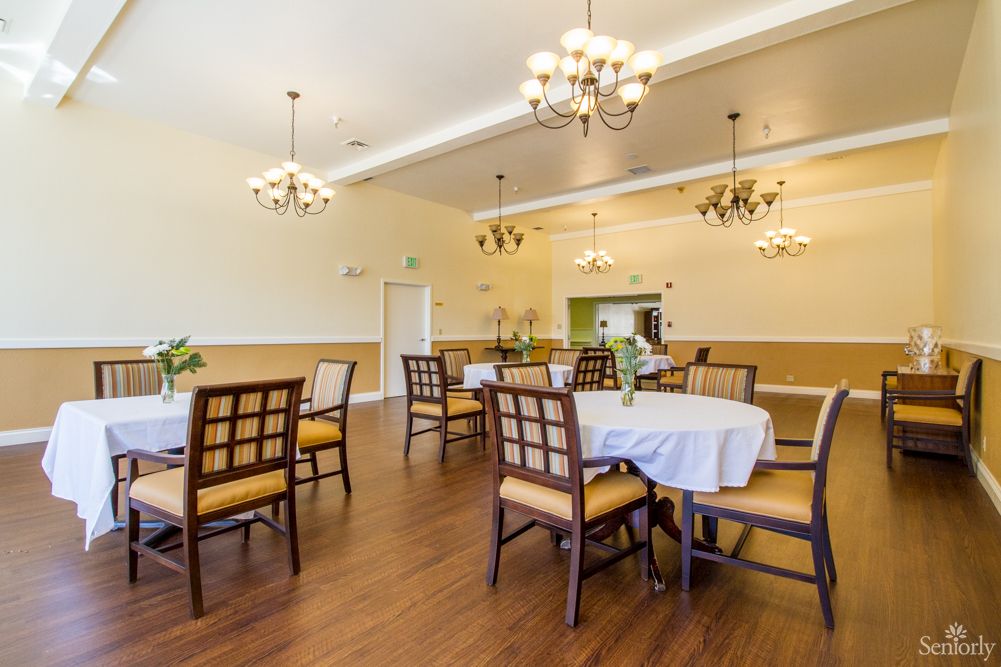 A well-lit dining room with elegantly set tables and chairs, featuring chandeliers and wooden flooring.