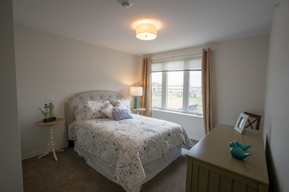 A cozy bedroom with a patterned bedspread and natural lighting from a large window.