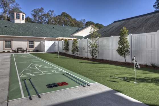 A backyard with a shuffleboard court, surrounded by a white fence and trees.