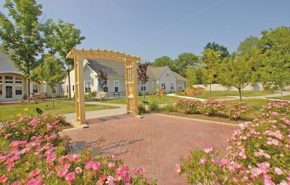 A picturesque garden with a wooden pergola, surrounded by blooming flowers and a paved pathway leading to a charming house.